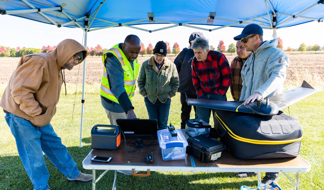 A group of GIS/Drone Technology students stand with their coordinator at an outdoor tent to look at information on a computer about the drone.