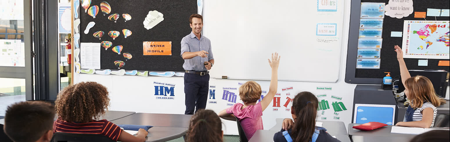 Elementary school teacher standing at the front of a classroom as students raise their hands to answer a question.