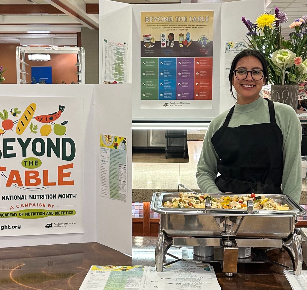 A student wearing an apron stands behind a table with food and posters at a wellness fair
