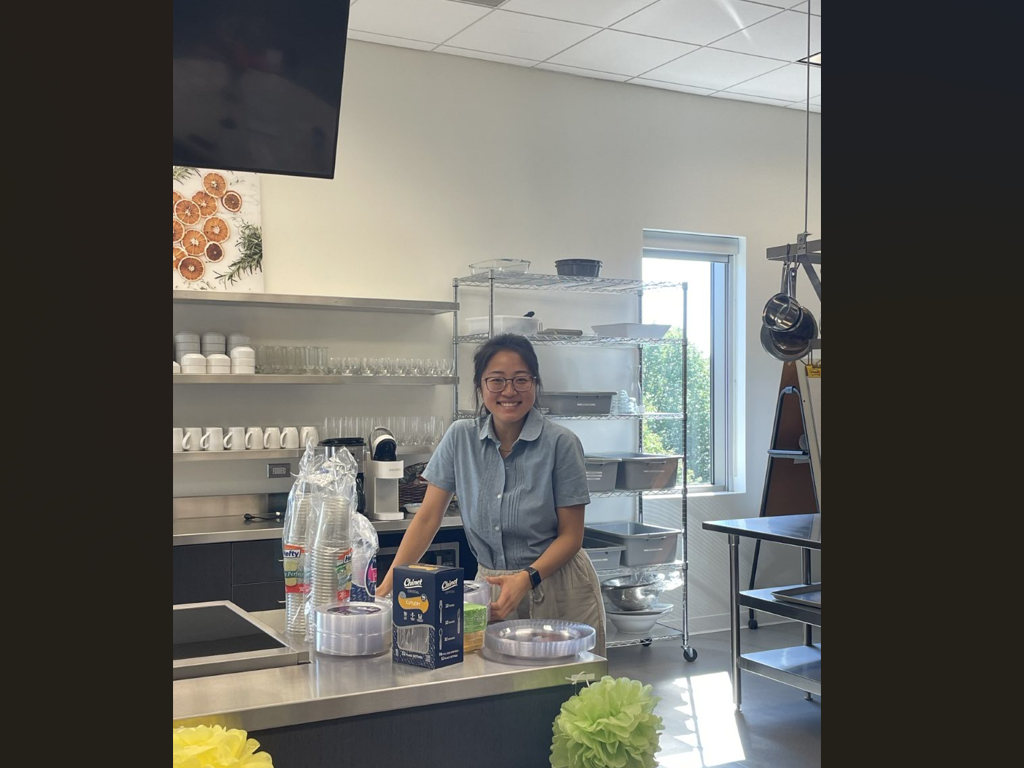 Dietetic internship student with glasses smiles in kitchen area, with cups and plates on a counter nearby