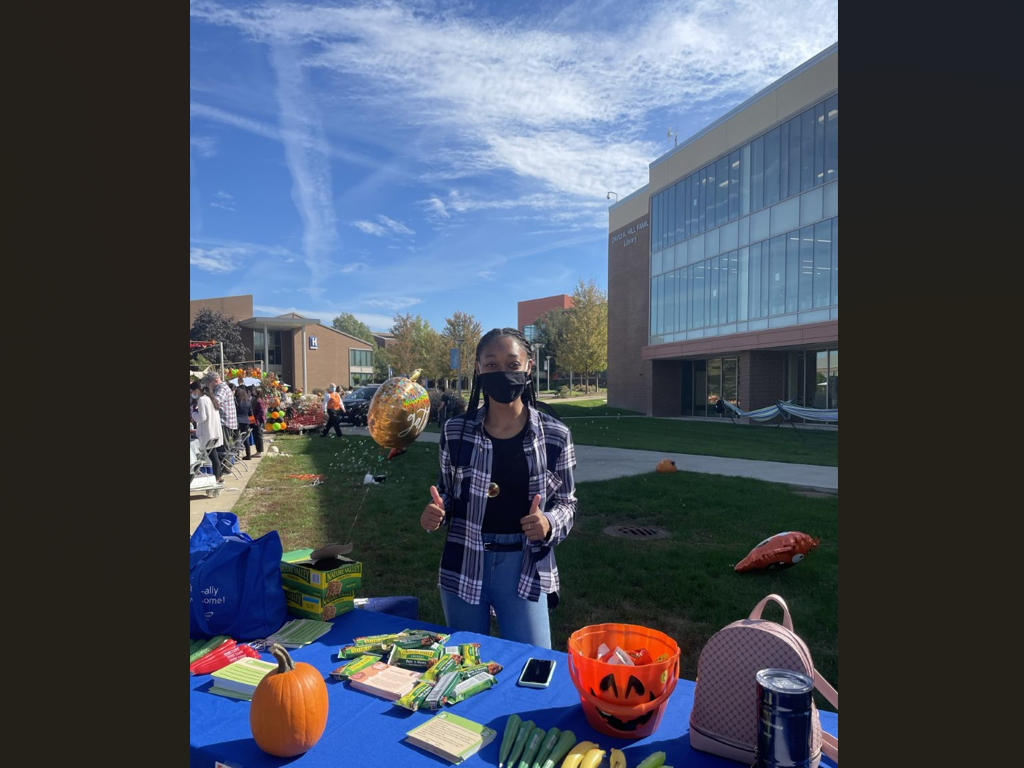Student gives a thumbs up at an outdoor tabling event with info and a Halloween pumpkin on the table.