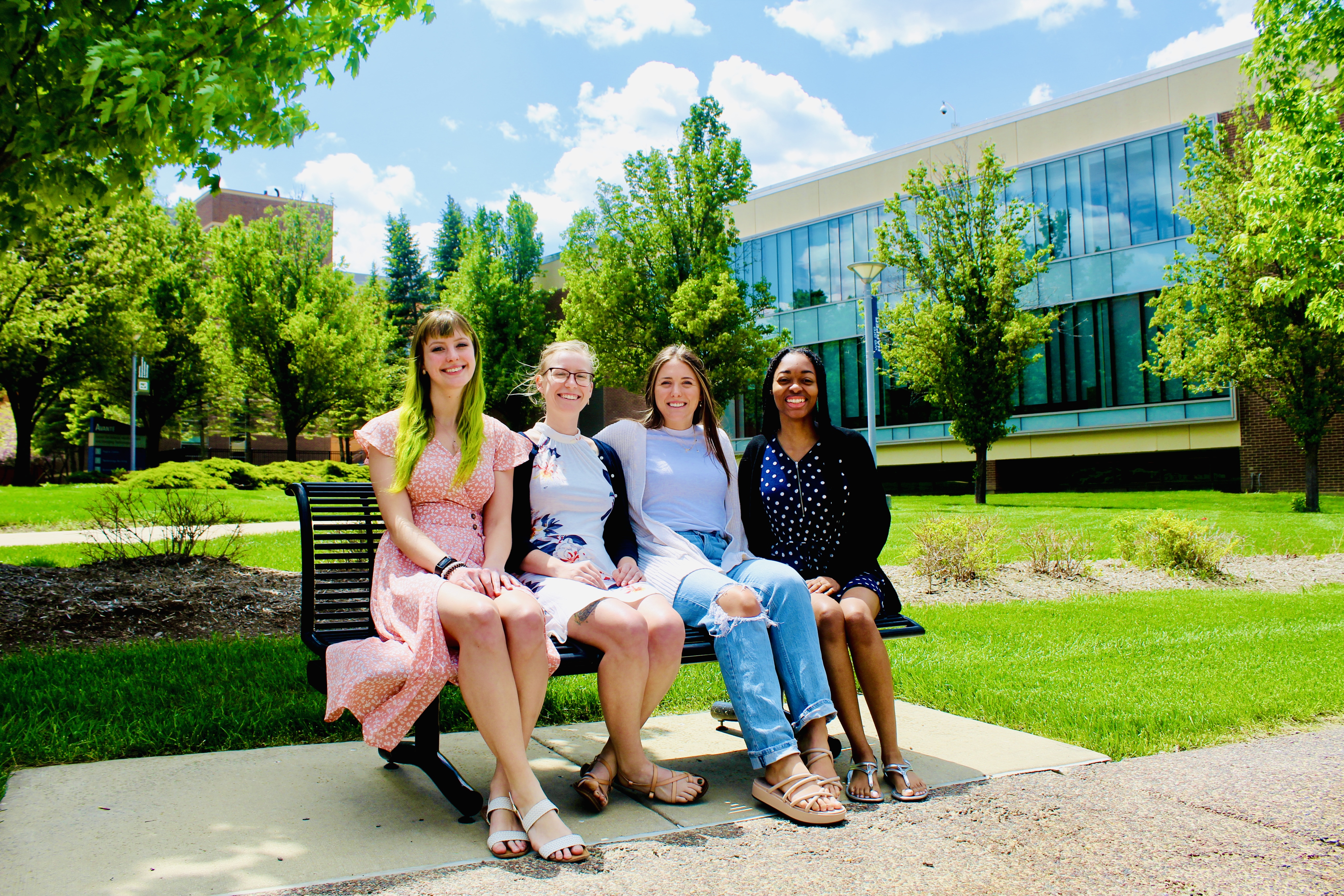 Four dietetic internship students sit together on an outdoor bench on a sunny day on campus.