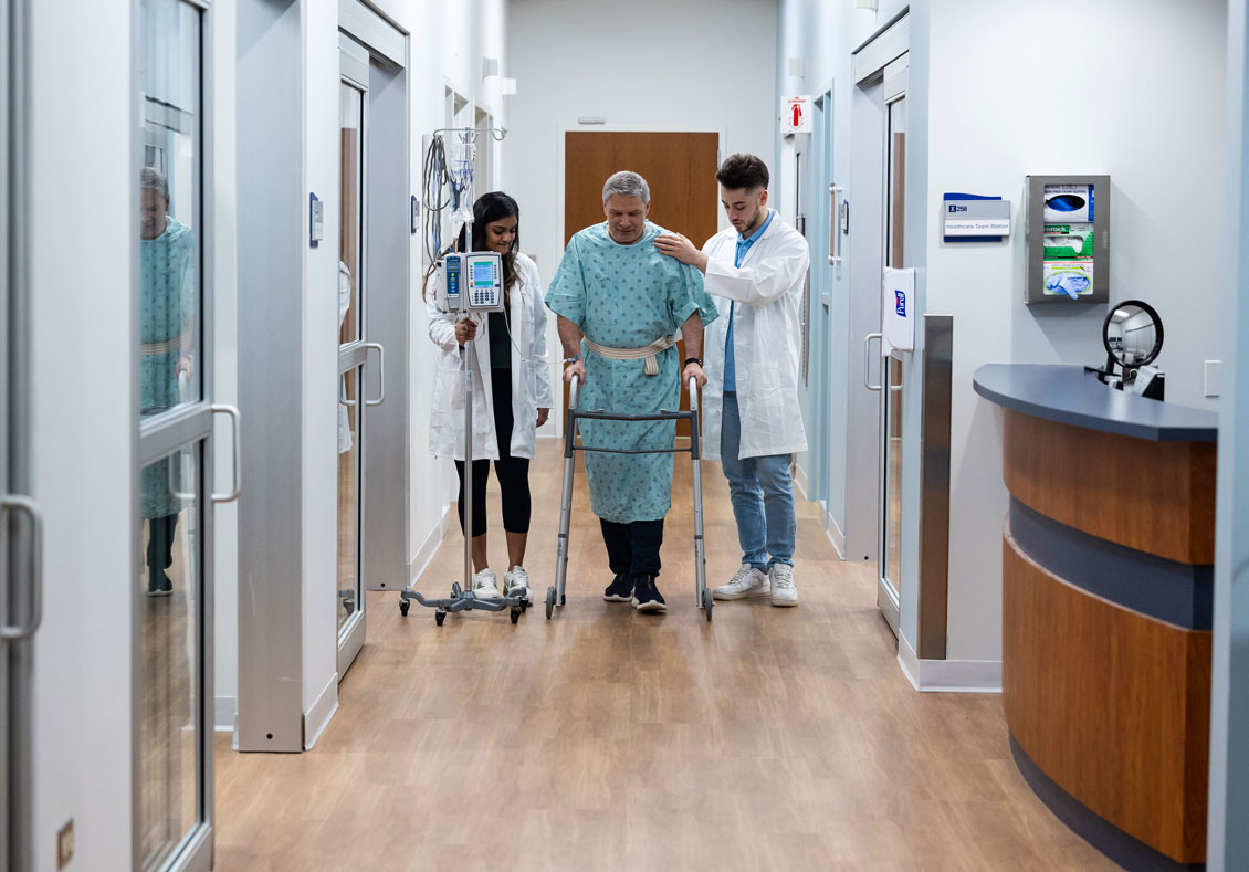 distant shot of two students helping a simulation patient walk down the hall