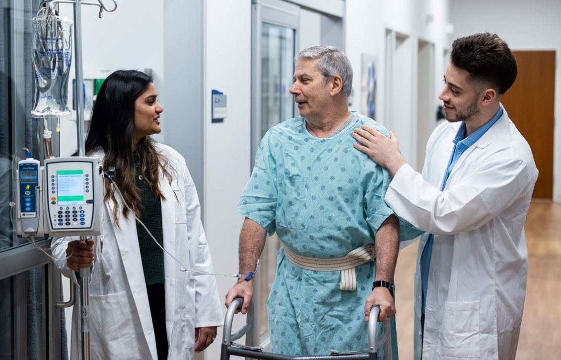 two students talk to a simulation patient in the hallway