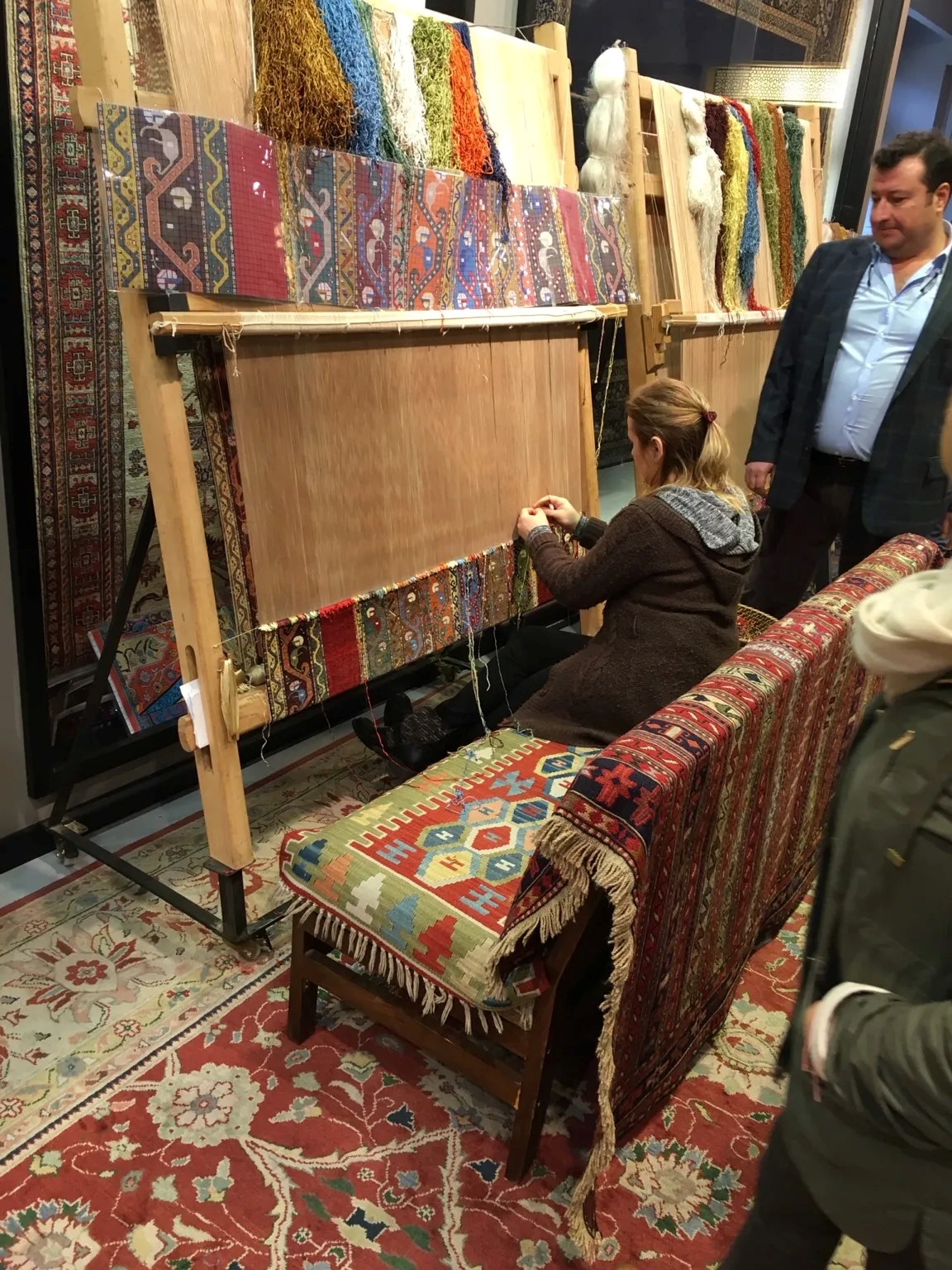 Woman sat weaving at a loom with multi-print carpets in the background