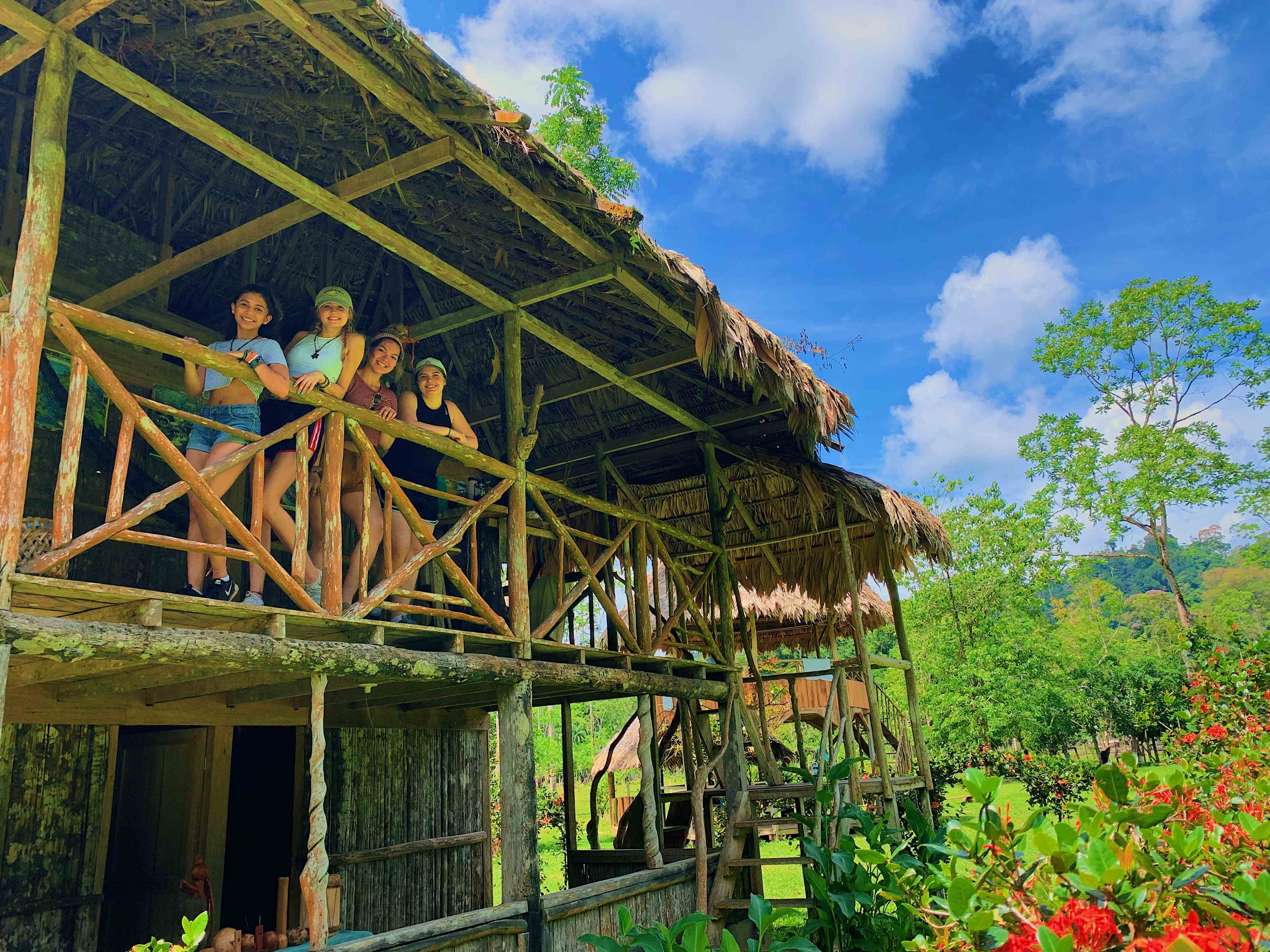 Three young women on a stilted, traditional home with lush forest in the background