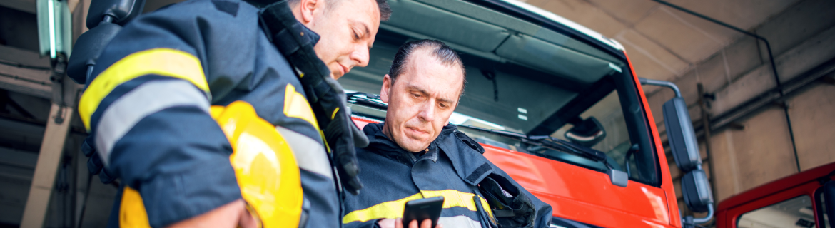 Two firefighters in full gear standing in front of a firetruck at the station garage.