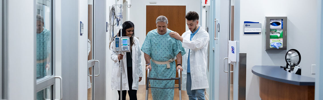 distant shot of two students helping a simulation patient walk down the hall