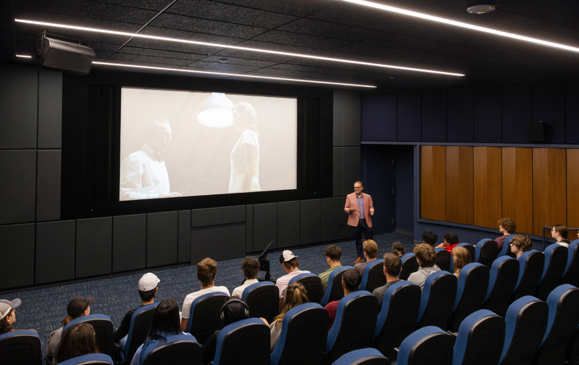 Film students fill the seats of the Movie Screening room. A movie is playing on the large projector screen while a professor lectures about the film.