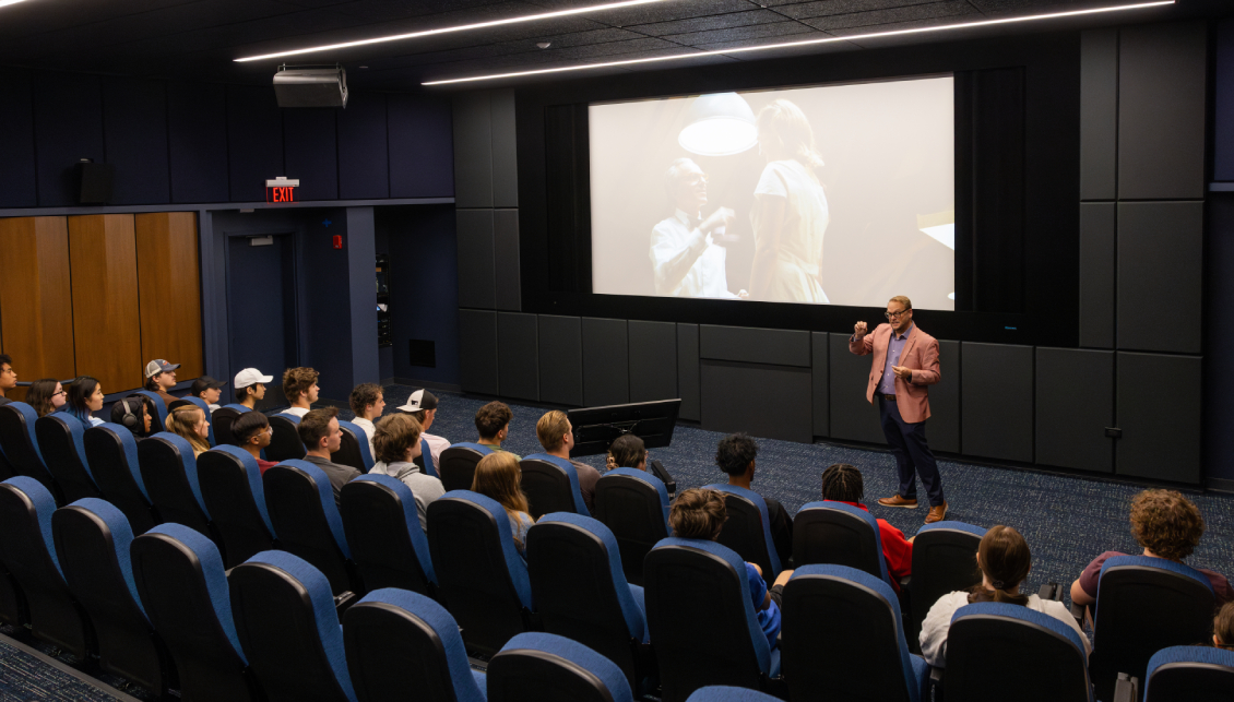 Film students fill the seats of the Movie Screening room. A movie is playing on the large projector screen while a professor lectures about the film, photographed at a closer angle.