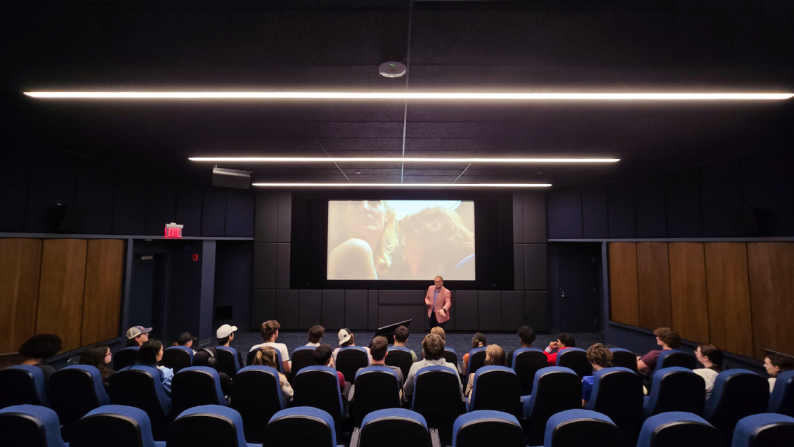 Students fill the first few rows of seats in the Movie Screening Room/Film Lab at Harper College while a professor lectures at the front, viewed from directly behind the seats that face the large projector screen