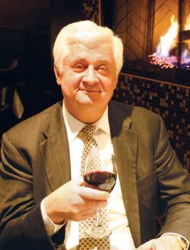 George Kalkounos wearing a gray suit, seated in front of a fire holding a glass of red wine.