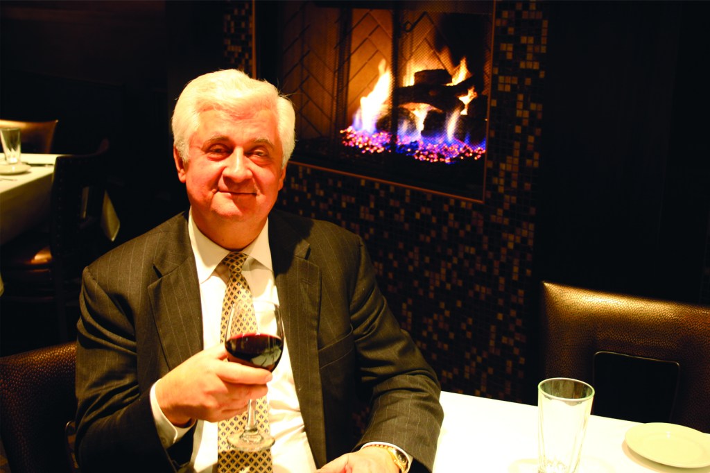 George Kalkounos sitting in front of a fire with a glass of wine.