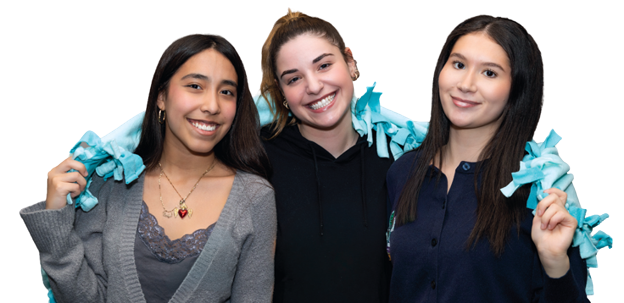 Three smiling Harper College students with a light blue blanket that they have made during the HOPE Day of Action around their shoulders.