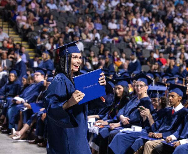 Graduates in blue caps and gowns sit at an outdoor ceremony, with a smiling woman in a kente stole at the center