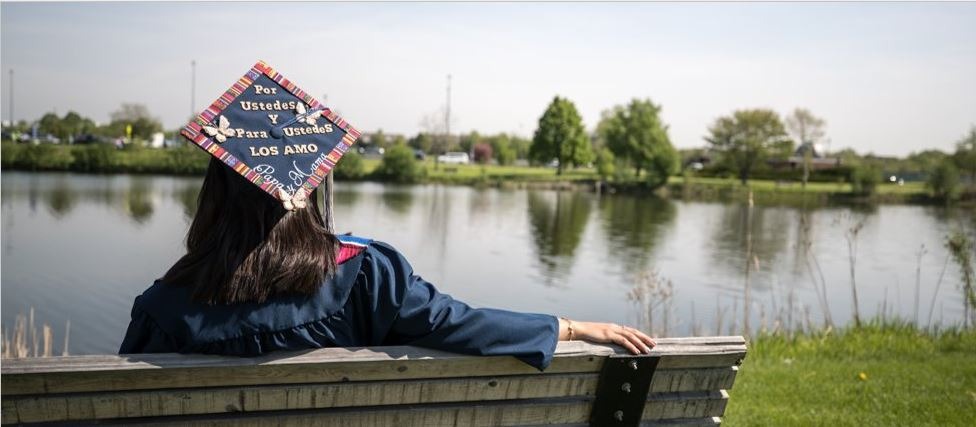 Image of Latinx Harper grad sitting on bench facing lake. Graduation cap has been decorated by hand to read "Por ustedes y para ustedes. Los amo, papa y mama