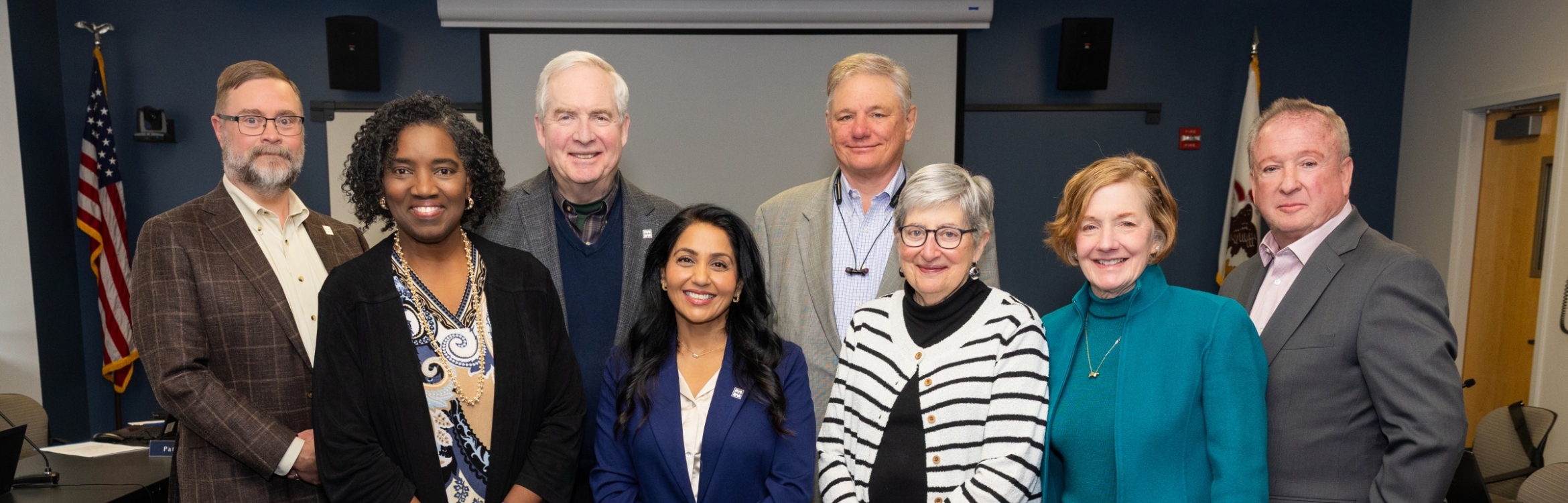 Group photo of eight board members standing in a conference room at Harper College.