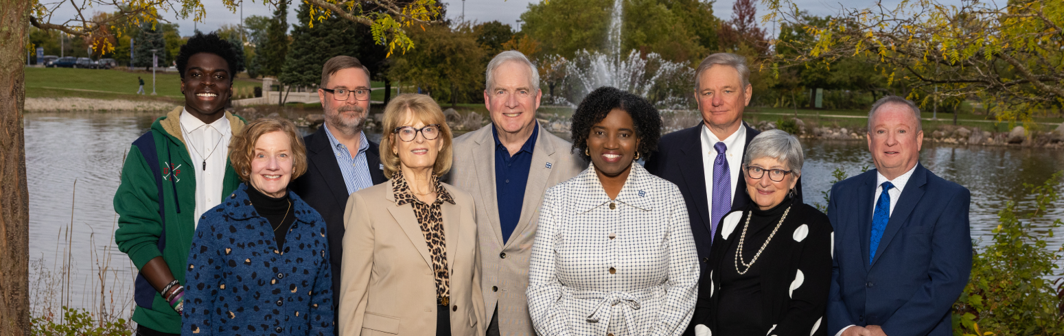 Group photo of nine board members standing outdoors at Harper College in front of a lake and fountain.