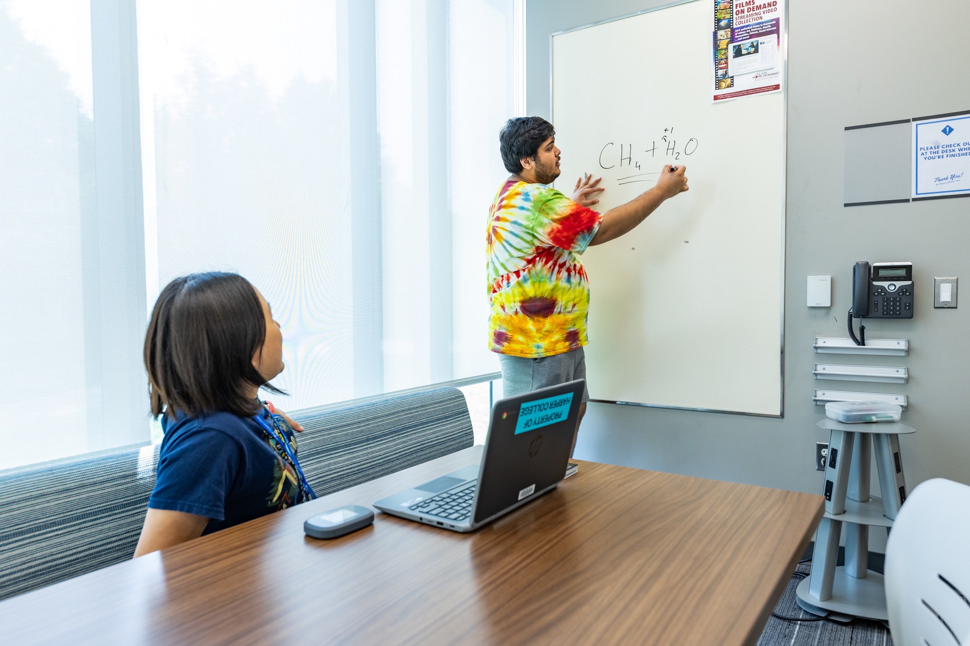 One standing student writes on a whiteboard while a student seated at a table looks on