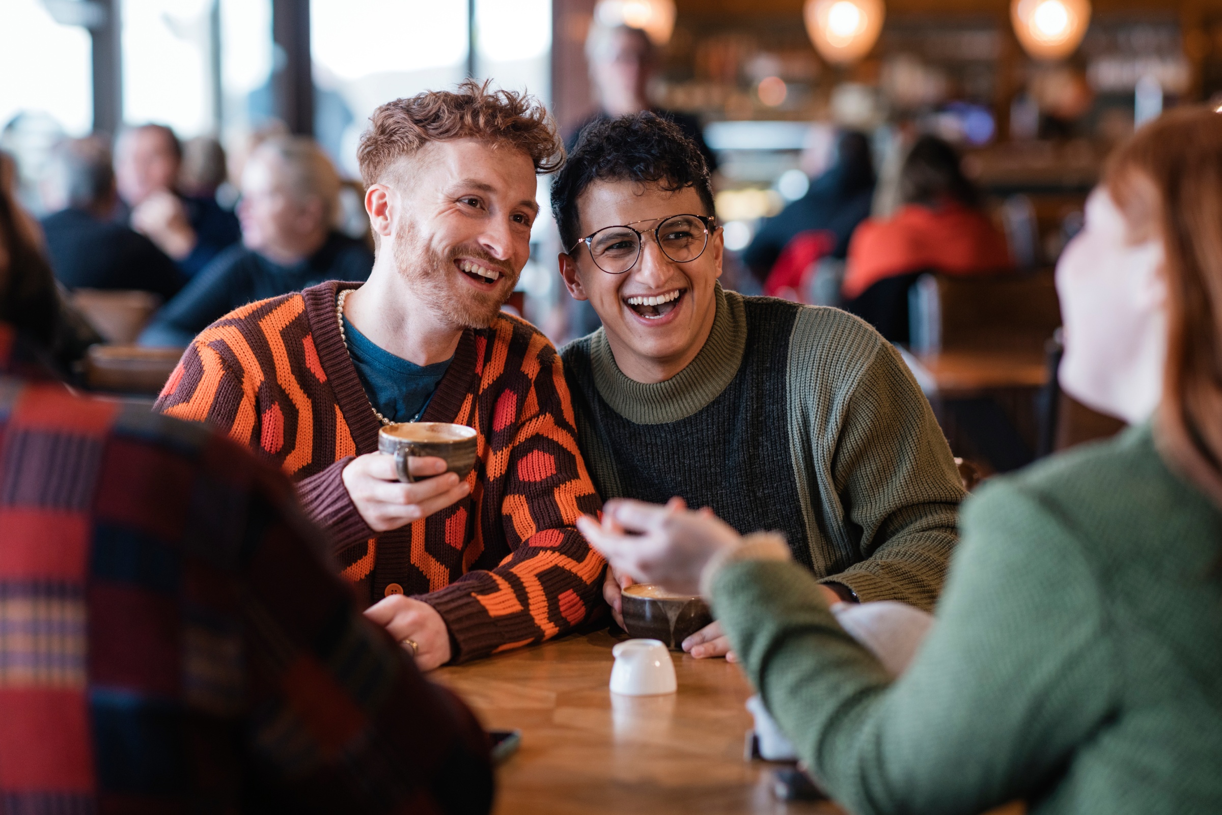 Two men smiling in a coffee shop and speaking with friends.
