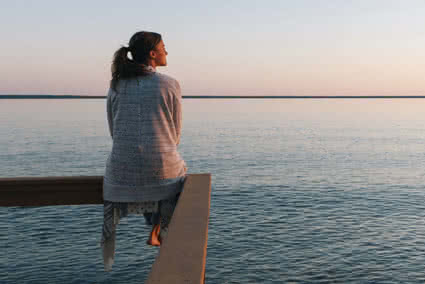 Young Woman sitting on the edge of a deck and looking out at the sea while the sun sets in the background