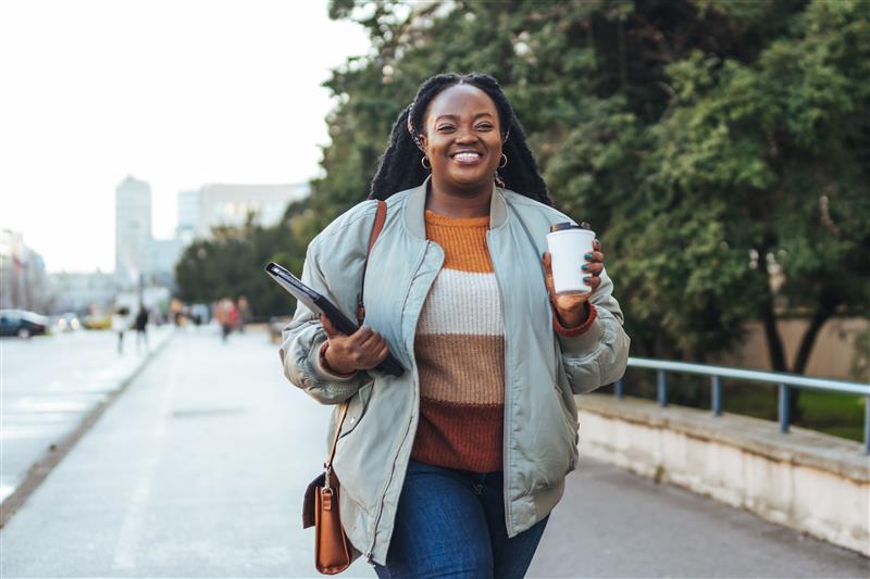 Young woman smiling with a coffee cup and laptop in her hands as she walks to work in the city.