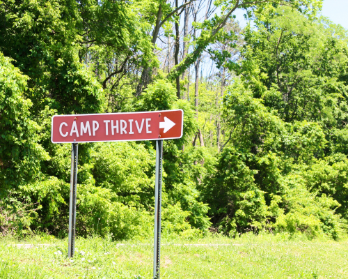 Photo of direction sign in woods, reading "Camp Thrive"
