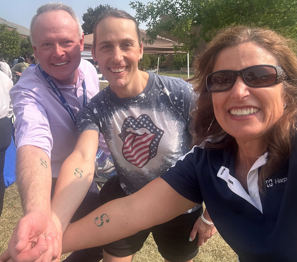 Harper College community members smiling at an outdoor campus event, showing temporary stamps on their arms