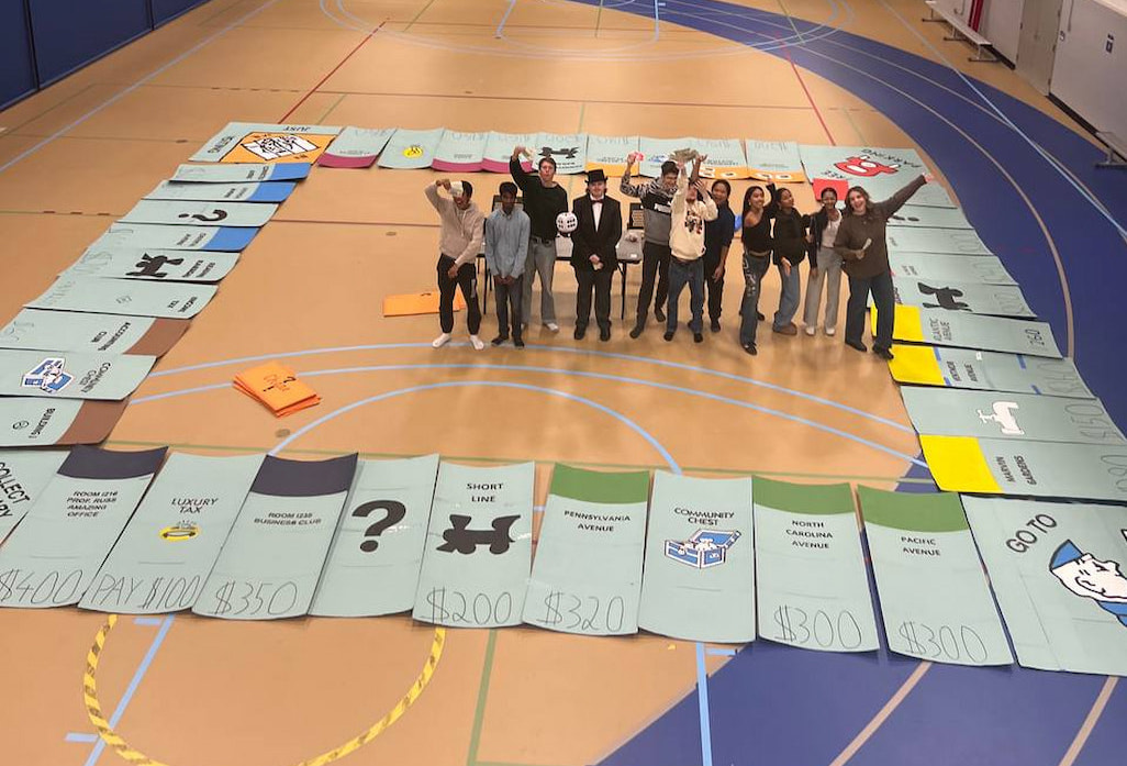 Harper College Business Club members standing on a gym floor around a large Monopoly-style board game during a group activity.