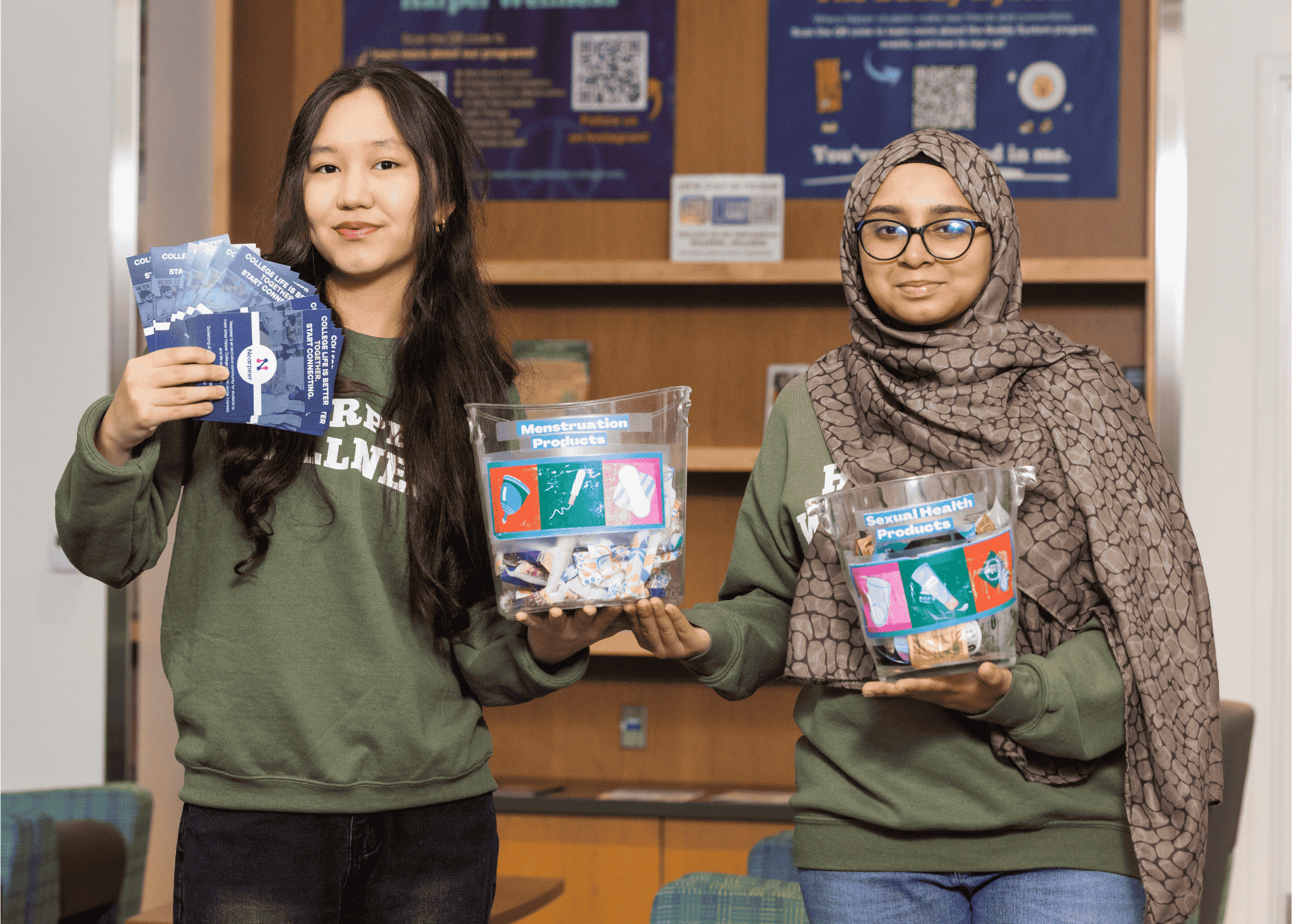 Two individuals wearing Harper Wellness sweatshirts stand indoors holding wellness materials. One holds several informational cards, and the other holds clear bins labeled “Menstruation Products” and “Sexual Health Products,” containing assorted supplies. A display with posters and QR codes is visible in the background.