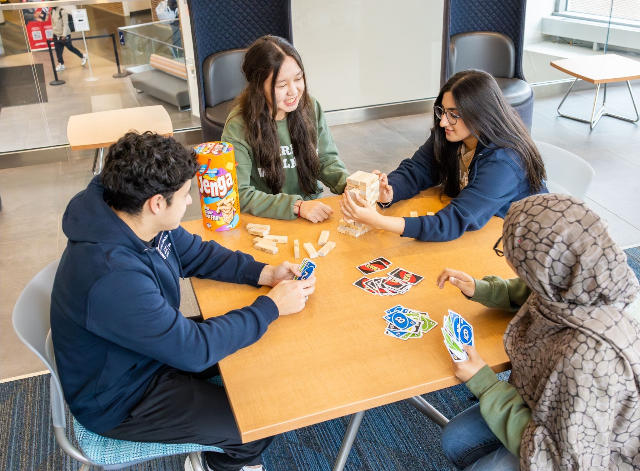 Five students sit around a table playing Jenga and card games, smiling and interacting in a casual indoor lounge setting.
