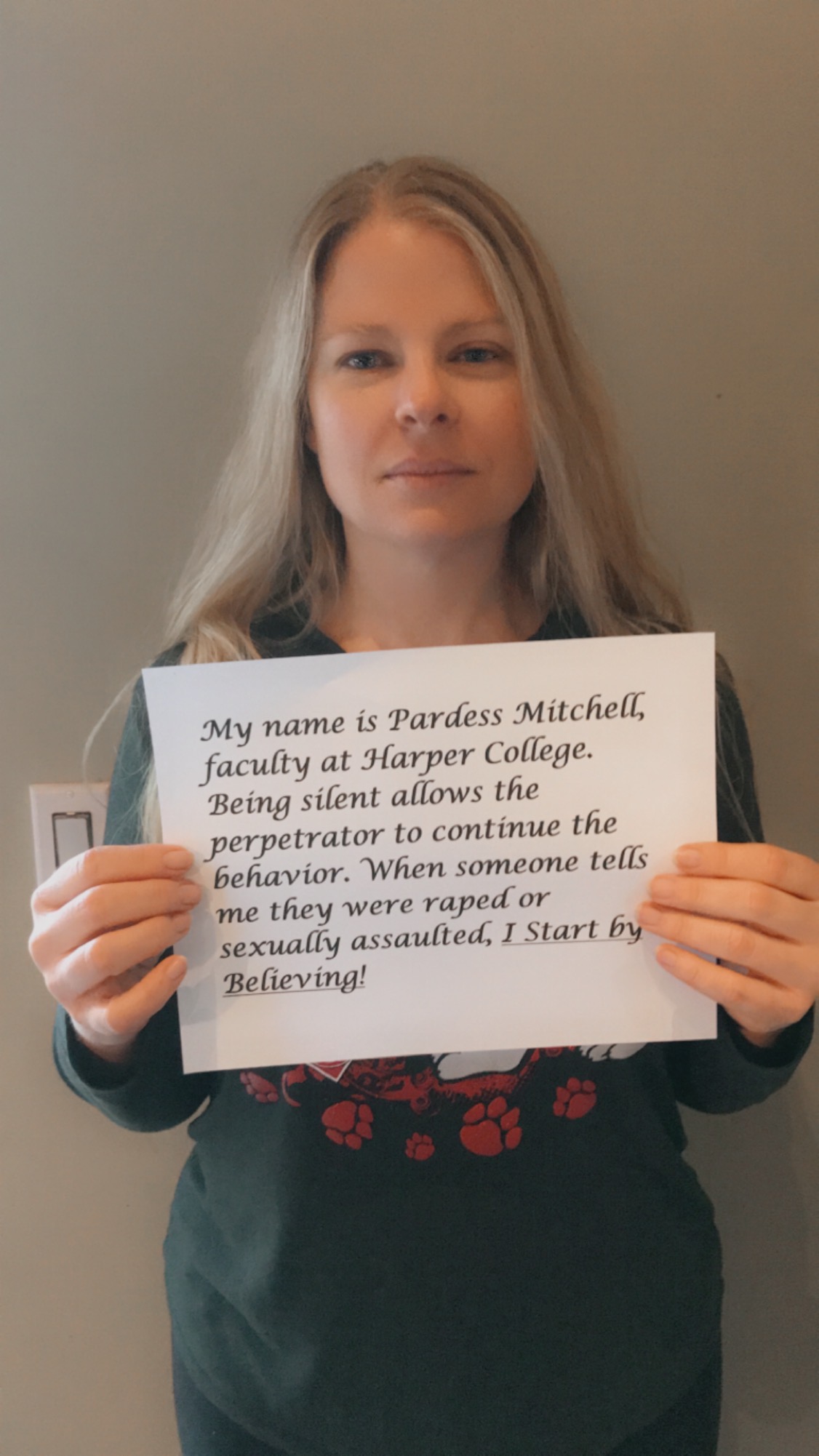 Photo of Pardess Mitchelle holding up a sign with her name on it and the words, "I start by believing." 