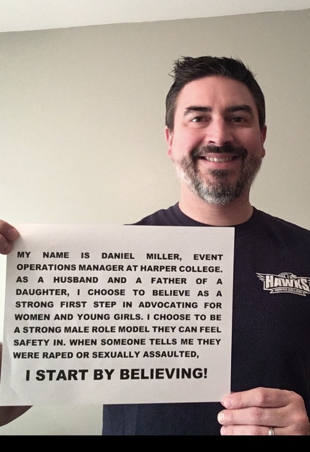 Photo of Daniel Miller holding up a sign with his name on it with the words, "I start by believing." 