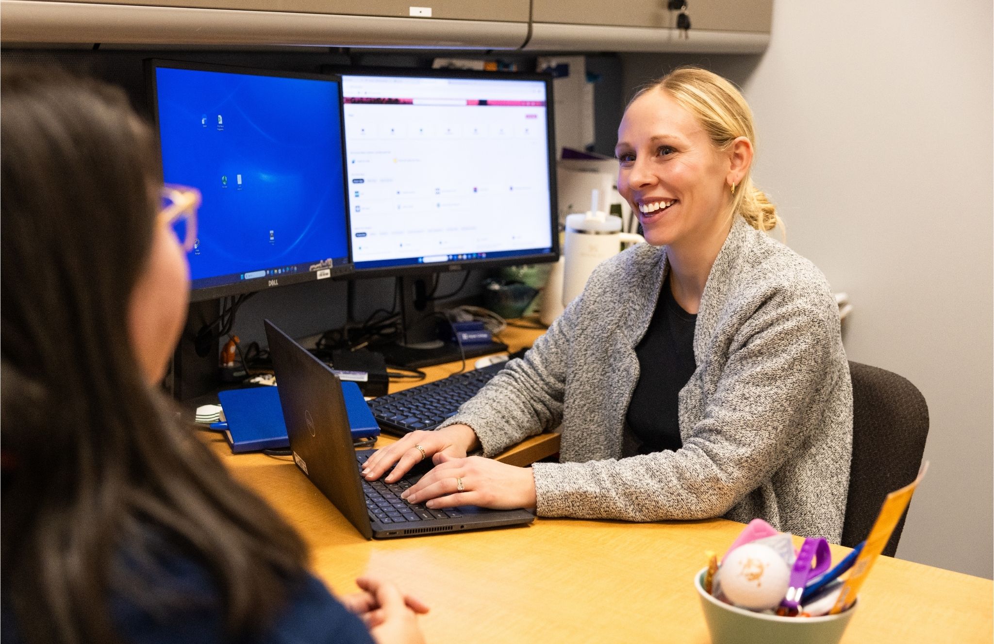 Two people sit across from each other at a desk in an office. One person is typing on a laptop, with two computer monitors displaying work screens behind them. The other person sits on the opposite side of the desk. Office supplies, including a cup holding various items, are on the desk.