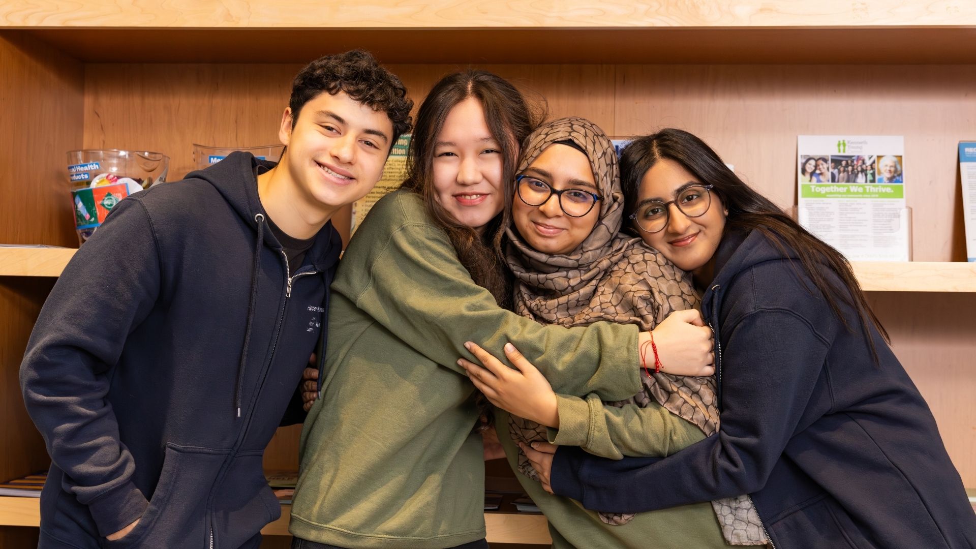 Four students stand close together in front of a bookshelf, smiling and hugging each other in a warm, supportive pose.