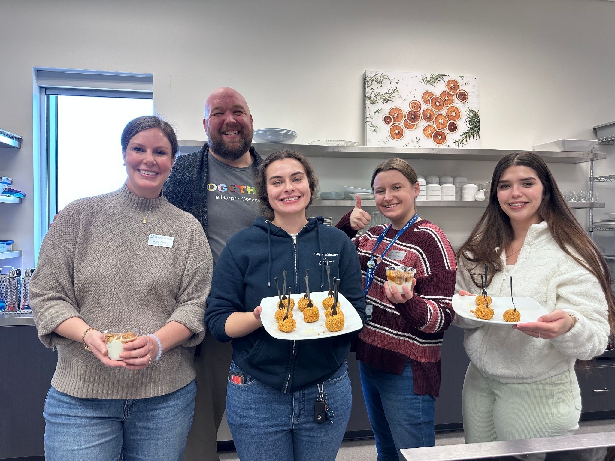 A group of five people standing together in a kitchen space, each holding plates or cups with small food items. Two individuals hold trays with several bite-sized snacks on sticks, while the others hold single servings in small cups. Behind them are shelves with dishes and a wall painting featuring orange slices.