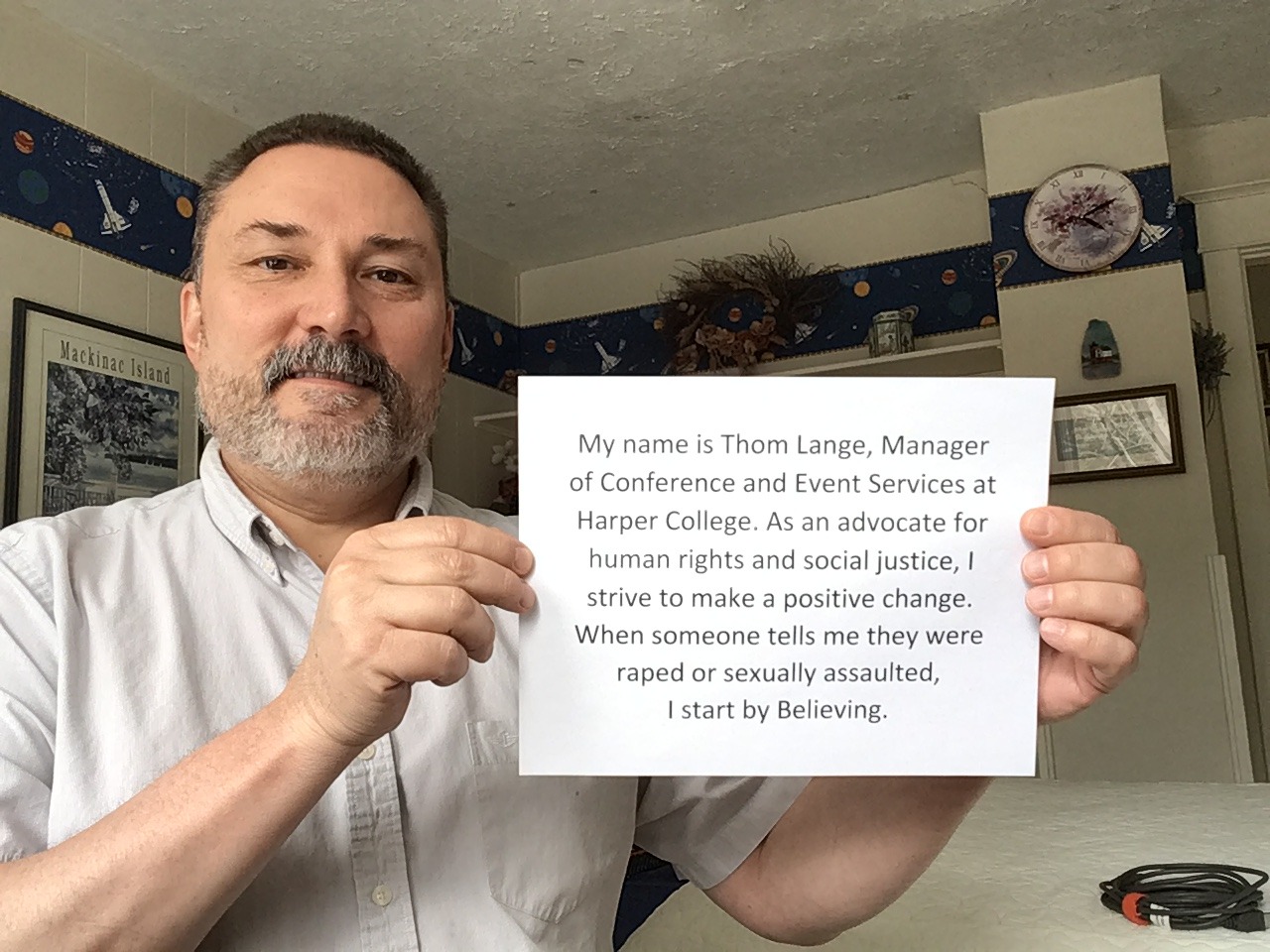 Photo of Thom Lange holding up a sign with his name on it and the words, "I start By Believing"