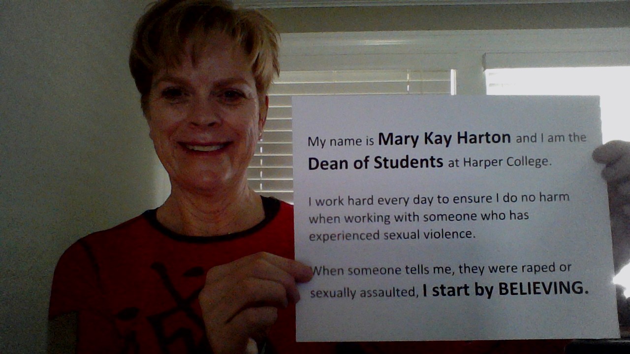 Photo of Mary Kay Harton holding up a sign that says her name and the words "I Start By Believing"